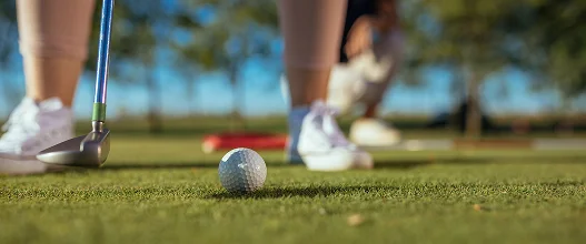 Jogador a preparar uma tacada num campo de minigolfe, com um taco e uma bola de golfe num green bem cuidado, durante um jogo ao ar livre.