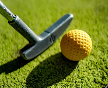 Grande plano de um taco de minigolfe colocado ao lado de uma bola de golfe amarela num green artificial, pronto para uma tacada de precisão.