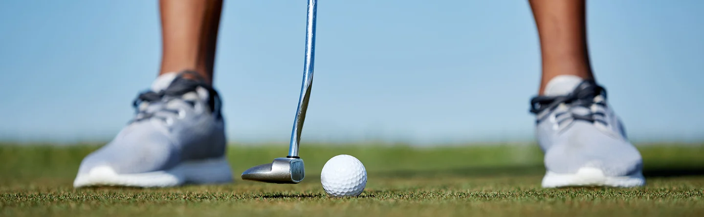 Imagem tirada de um ângulo baixo de um taco de golfe e de uma bola branca, alinhados para uma tacada num putting green exterior liso, com o jogador em pé atrás deles.
