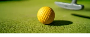 Bola de minigolfe amarela pousada num green artificial, com um taco de golfe ao fundo, destacando a precisão e a exatidão.