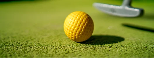 Bola de minigolfe amarela pousada num green artificial, com um taco de golfe ao fundo, destacando a precisão e a exatidão.